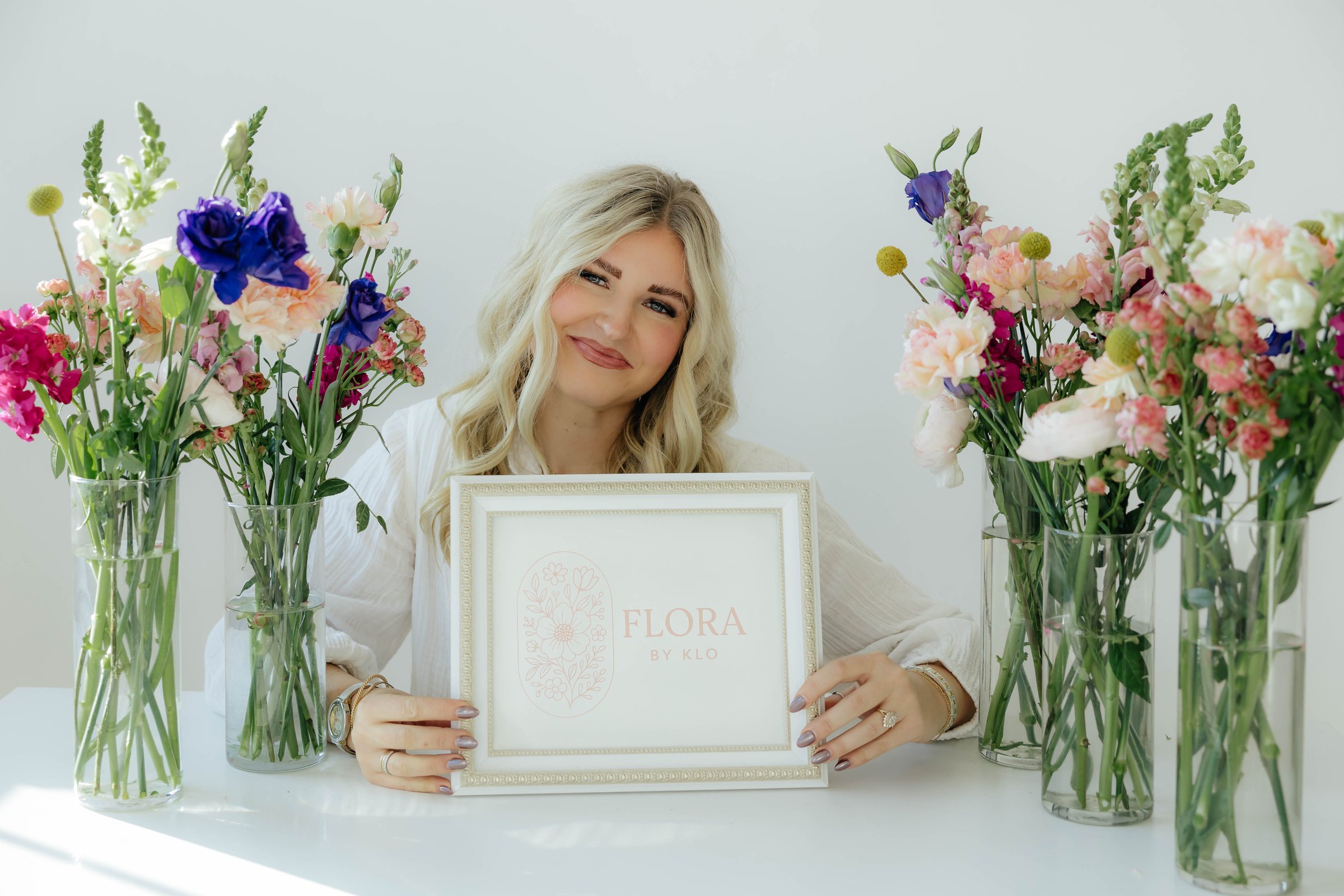 Kloe Brown holding the Flora by Klo sign between vases of fresh flowers